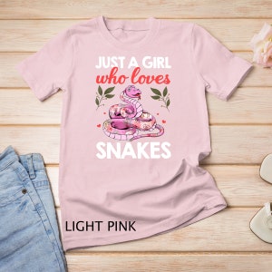 Könnte beinhalten: Ein hellrosa T-Shirt mit dem Text "Just a girl who loves snakes" und einer rosa Schlange mit Blumenmustern.
