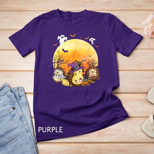 Pode incluir: Camiseta roxa com um design de Halloween. O desenho inclui três ouriços de desenho animado fantasiados, uma lua cheia, morcegos e fantasmas. A palavra "PURPLE" está impressa na parte inferior da camiseta.