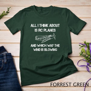 Könnte beinhalten: Ein dunkelgrünes T-Shirt mit weißem Text, der "ALL I THINK ABOUT IS RC PLANES AND WHICH WAY THE WIND IS BLOWING" lautet. Über dem Text befindet sich eine weiße Silhouette eines Flugzeugs. Der Text "FORREST GREEN" befindet sich am unteren Rand des Bildes.
