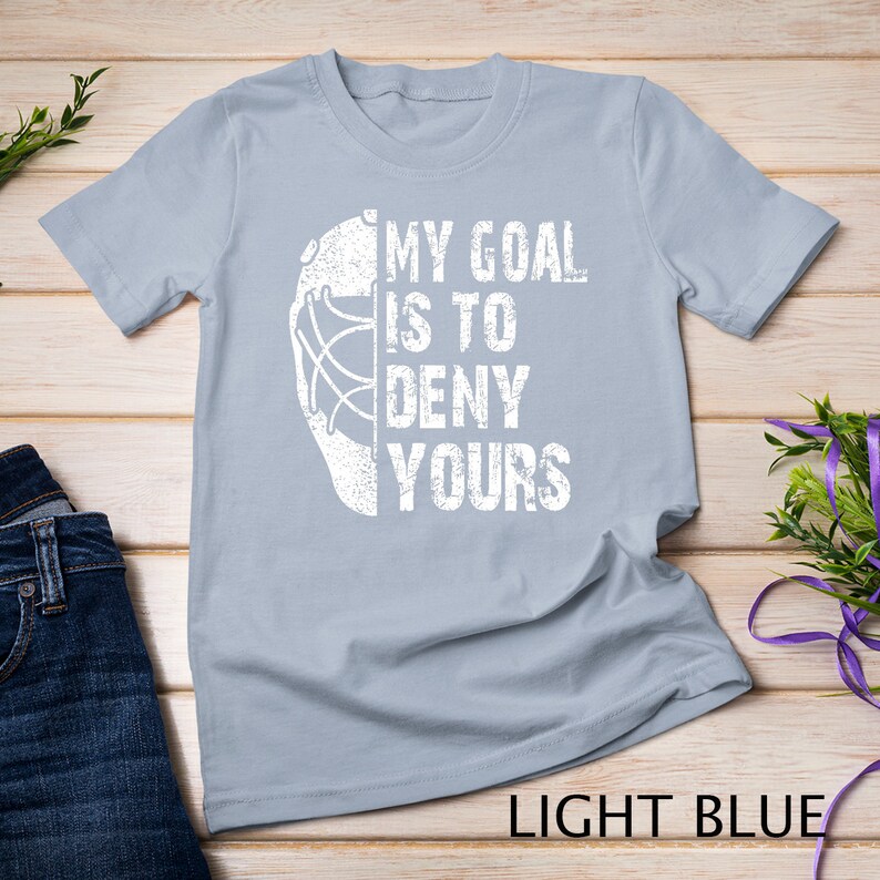 May include: Light blue t-shirt featuring a white hockey goalie mask graphic and the text "My goal is to deny yours".