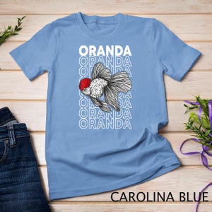 May include: A light blue t-shirt with a white graphic of an oranda goldfish. The text "ORANDA" is repeated in white letters down the front of the shirt.