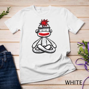 May include: White t-shirt with a cartoon illustration of a sock monkey meditating. The monkey has red hair, a white face, and red lips. The monkey is wearing a white sock with a red stripe.