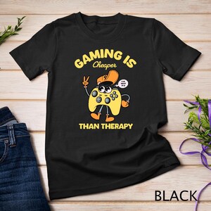 May include: Black t-shirt with a yellow and black video game controller graphic. The text "Gaming is Cheaper Than Therapy" is printed on the shirt.