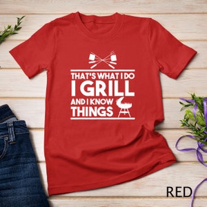 May include: Red t-shirt with white text that reads "That's what I do I grill and I know things". The text is surrounded by a grill and crossed grilling tools.