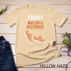 Op de afbeelding: Een beige T-shirt met de tekst "FIGHT MULTIPLE SCLEROSIS" en twee oranje hardloopschoenen gemaakt van oranje bewustzijnslintjes. De tekst op de schoenen luidt "WALK PROUD" en "WALK FOR A CURE".