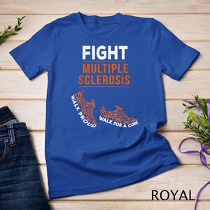 Op de afbeelding: Koninklijk blauw T-shirt met de tekst "FIGHT MULTIPLE SCLEROSIS" in wit. Het shirt heeft twee oranje hardloopschoenen gemaakt van oranje bewustzijnslintjes. De tekst "WALK PROUD" staat op de linker schoen en "WALK FOR A CURE" staat op de rechter schoen.