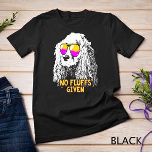 May include: Black t-shirt featuring a white dog wearing pink and yellow sunglasses. The text "NO FLUFFS GIVEN" is printed in yellow and pink on the shirt.