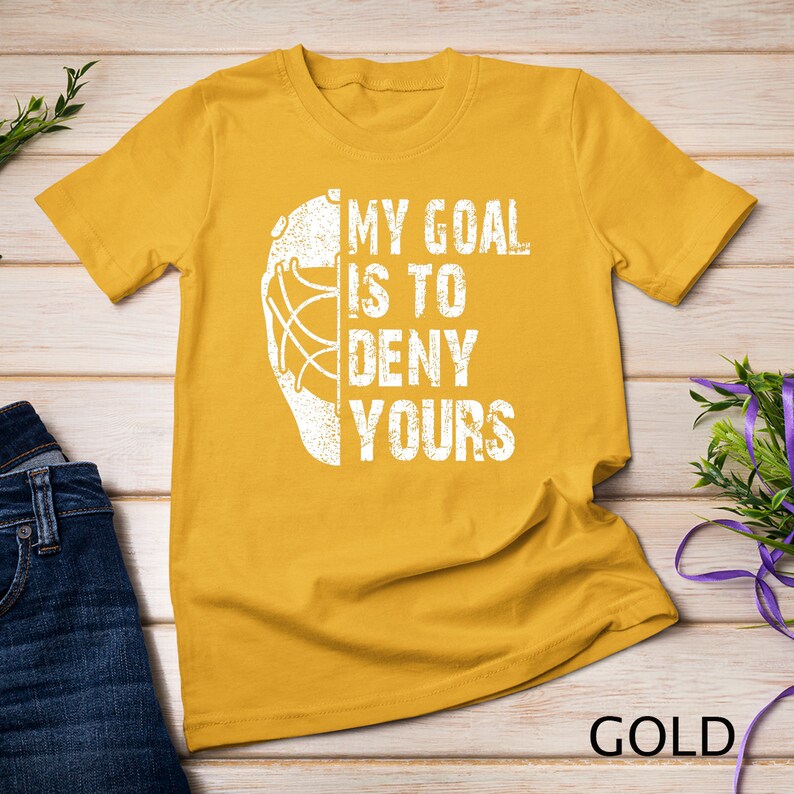 May include: A gold t-shirt featuring a white hockey mask graphic and the text "My goal is to deny yours".