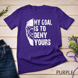 May include: Purple t-shirt featuring a white hockey goalie mask graphic and the text "My goal is to deny yours".