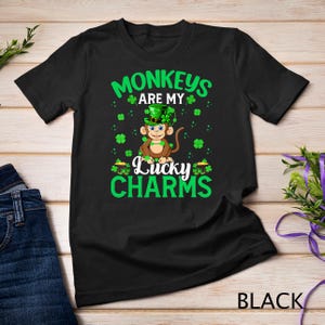 Può includere: Maglietta nera con un design a trifoglio verde e il testo "Monkeys are my Lucky Charms". Una scimmia di cartone animato con un cappello verde è al centro del design.