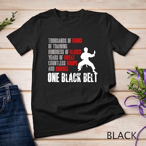 Puede incluir: Camiseta negra con texto blanco que dice "Miles de horas de entrenamiento, cientos de clases, años de sudor, innumerables golpes y moretones, un cinturón negro". Una silueta blanca de una persona en una pose de karate se muestra sobre el texto.