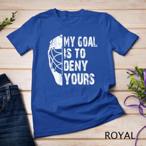 May include: A royal blue t-shirt featuring a white hockey goalie mask and the text "My goal is to deny yours".
