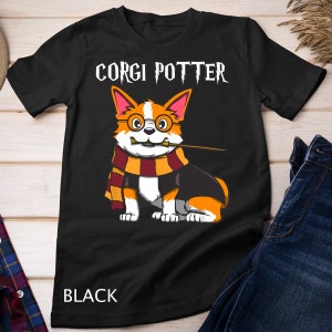 Puede incluir: Camiseta negra con una imagen de dibujos animados de un corgi con una bufanda de Hogwarts y gafas. El texto "Corgi Potter" está impreso en la camiseta.