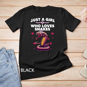 Op de afbeelding: Zwart T-shirt met de tekst "Just a girl who loves snakes" en een roze slang met een bloem op zijn kop. Er zijn roze harten rond de slang.