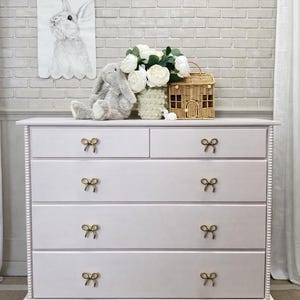 Pode incluir: Uma cômoda rosa claro com seis gavetas e puxadores dourados em forma de laço. A cômoda tem uma base decorativa e uma borda com contas. Um coelho de pelúcia, flores e uma cesta tecida em forma de casa estão em cima.