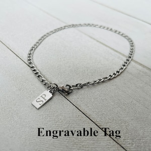 Peut inclure: Un bracelet en chaîne argentée avec une petite étiquette rectangulaire gravée des lettres "SP". L'étiquette est attachée à la chaîne par un fermoir à mousqueton. Le bracelet est sur une surface en bois blanc.  Étiquette gravable