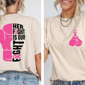 Pode incluir: Camiseta bege com um gr&aacute;fico de luva de boxe rosa e o texto "HER FIGHT IS OUR FIGHT" nas costas. A frente da camisa apresenta um par de luvas de boxe rosa. A camisa &eacute; feita de um material macio.