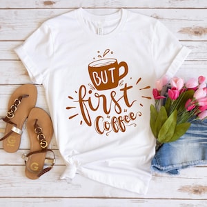 May include: White t-shirt with the phrase "But first, Coffee" in brown script and a coffee cup graphic. The shirt is styled with sandals and flowers on a white wooden surface.