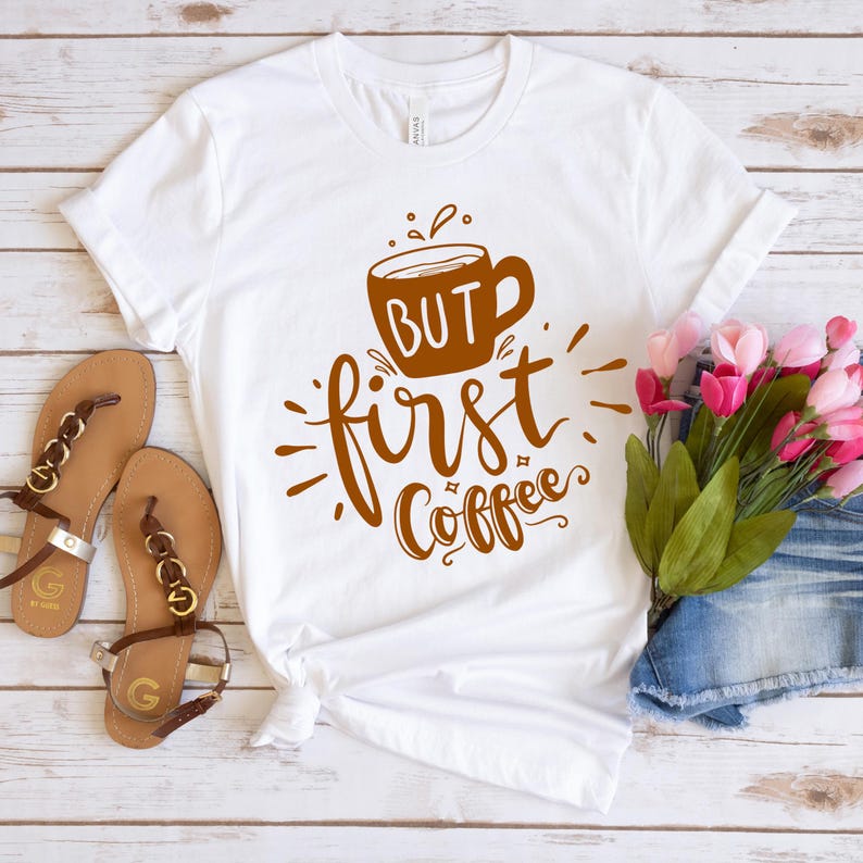 May include: White t-shirt with the phrase "But first, Coffee" in brown script and a coffee cup graphic. The shirt is styled with sandals and flowers on a white wooden surface.