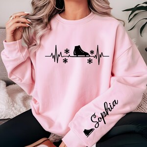 Op de afbeelding: Lichtroze sweatshirt met een zwarte afbeelding van een schaats, sneeuwvlokken en een hartslaglijn. De naam "Sophia" staat in cursief op de mouw. Onder de naam staat een klein icoon van een schaats.