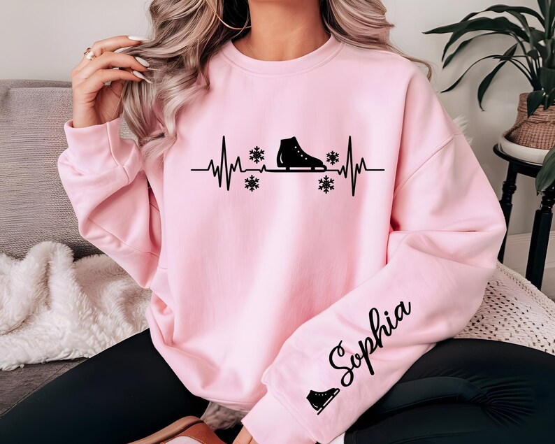 Op de afbeelding: Lichtroze sweatshirt met een zwarte afbeelding van een schaats, sneeuwvlokken en een hartslaglijn. De naam "Sophia" staat in cursief op de mouw. Onder de naam staat een klein icoon van een schaats.