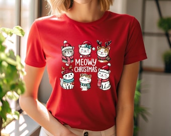 May include: Red t-shirt featuring a graphic of six cartoon cats wearing festive winter hats and scarves. The text "MEOWY CHRISTMAS" is printed in white. The cats are in various poses, adding a playful touch to the holiday theme.