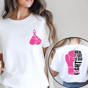 Pode incluir: Camiseta branca com um design de luva de boxe e fita rosa. A frente da camiseta apresenta duas luvas de boxe rosa com uma fita rosa. A parte de tr&aacute;s da camiseta tem uma luva de boxe rosa com o texto "HER FIGHT IS OUR FIGHT".