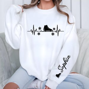 Op de afbeelding: Witte sweatshirt met een zwarte afbeelding van een schaats in een hartslaglijn, met sneeuwvlokken. De naam "Sophia" staat in zwart schrift op de mouw. Een kleine schaatsafbeelding staat eronder.