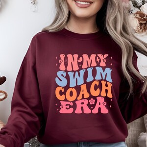May include: Burgundy sweatshirt with the text "IN MY SWIM COACH ERA" in a retro font. The text is in shades of pink, orange, and blue. The sweatshirt has a crew neck and long sleeves.