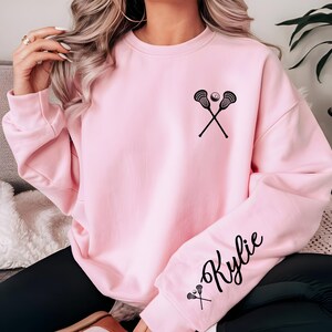 May include: A light pink sweatshirt featuring a black lacrosse stick and ball graphic on the chest. The right sleeve displays the name "Kylie" in a script font, along with a small lacrosse stick graphic. The sweatshirt has a crew neck and long sleeves.