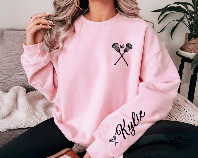May include: A light pink sweatshirt featuring a black lacrosse stick and ball graphic on the chest. The right sleeve displays the name "Kylie" in a script font, along with a small lacrosse stick graphic. The sweatshirt has a crew neck and long sleeves.