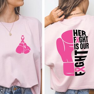 Pode incluir: T-shirt rosa claro com um design de luva de boxe rosa e uma fita rosa na frente. A parte de tr&aacute;s da camisa exibe uma luva de boxe rosa e o texto "HER FIGHT IS OUR FIGHT" em preto.
