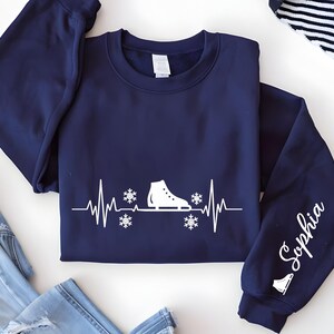 Op de afbeelding: Marineblauwe sweatshirt met een witte afbeelding van een schaats, sneeuwvlokken en een hartslaglijn. De mouw toont de naam "Sophia" in witte letters. Een winterkledingstuk.