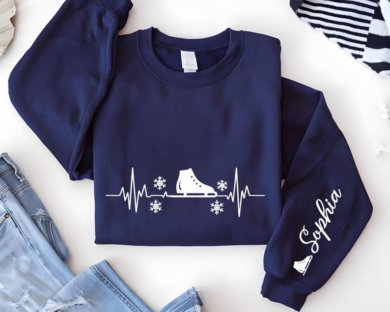 Op de afbeelding: Marineblauwe sweatshirt met een witte afbeelding van een schaats, sneeuwvlokken en een hartslaglijn. De mouw toont de naam "Sophia" in witte letters. Een winterkledingstuk.