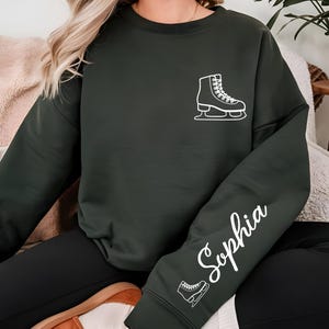 May include: Dark green sweatshirt featuring a white ice skate graphic on the chest and the name "Sophia" in cursive on the sleeve. The sweatshirt is a solid color and has a relaxed fit, suitable for casual wear.