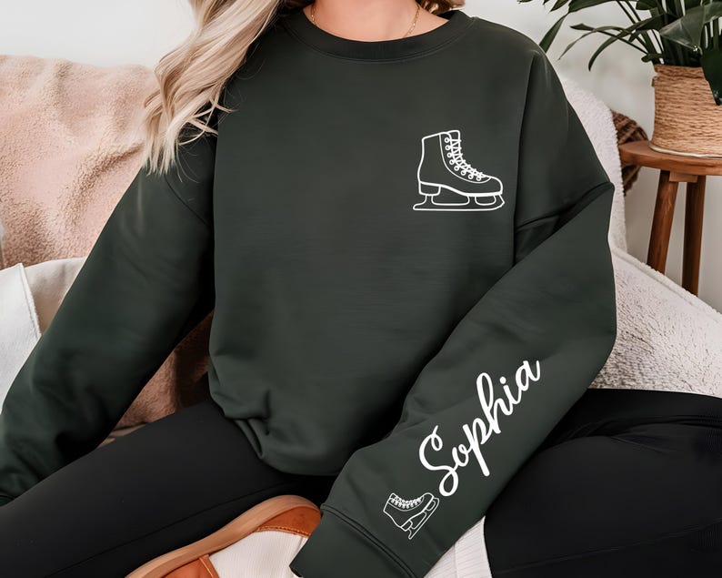 May include: Dark green sweatshirt featuring a white ice skate graphic on the chest and the name "Sophia" in cursive on the sleeve. The sweatshirt is a solid color and has a relaxed fit, suitable for casual wear.