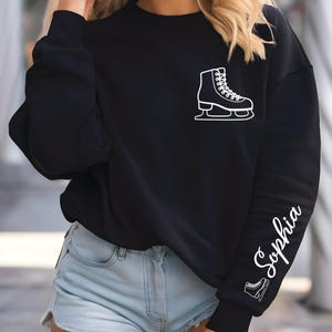 May include: Black sweatshirt with a white ice skate graphic on the chest and the name "Sophia" in cursive script down the left sleeve. A smaller ice skate graphic is below the name. The sweatshirt is a casual style.