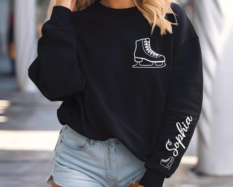 May include: Black sweatshirt with a white ice skate graphic on the chest and the name "Sophia" in cursive script down the left sleeve. A smaller ice skate graphic is below the name. The sweatshirt is a casual style.