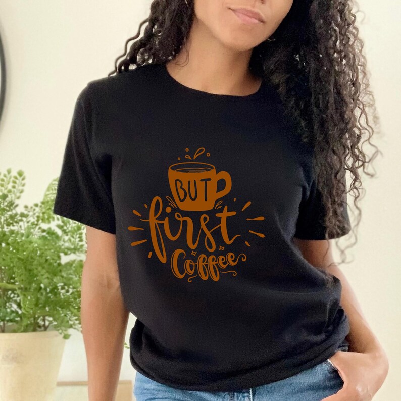 May include: Black t-shirt with the phrase "BUT first Coffee" in a brown, stylized font. The design includes a coffee cup illustration. The shirt is a crew neck and short-sleeved.