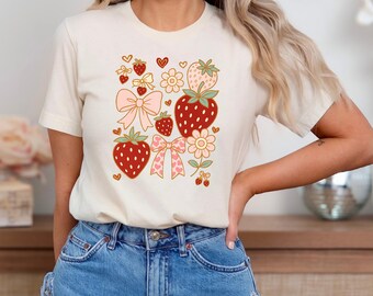 May include: A white t-shirt with a graphic design of strawberries, bows, and flowers in shades of red, pink, and green.