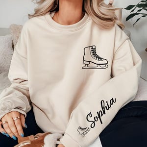 May include: Cream-colored sweatshirt with a black outline of an ice skate on the chest. The name "Sophia" is written in cursive on the sleeve, along with a small ice skate graphic. The sweatshirt is a casual style.