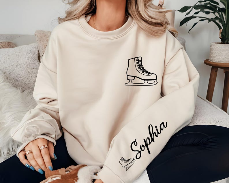 May include: Cream-colored sweatshirt with a black outline of an ice skate on the chest. The name "Sophia" is written in cursive on the sleeve, along with a small ice skate graphic. The sweatshirt is a casual style.