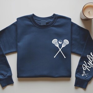 May include: Navy blue sweatshirt featuring a white lacrosse stick and ball graphic on the chest. The right sleeve displays the name "Kylie" in a cursive font, along with a small lacrosse stick graphic. A candle sits in the upper right corner.