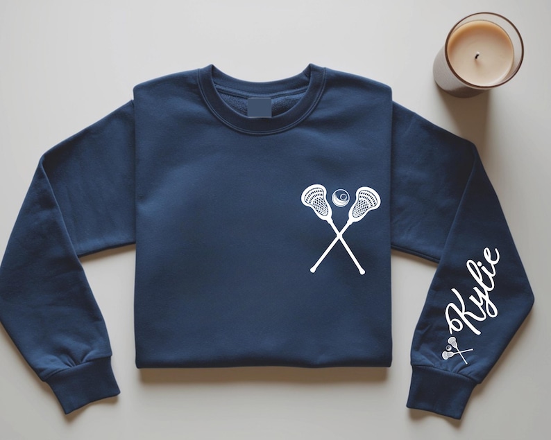 May include: Navy blue sweatshirt featuring a white lacrosse stick and ball graphic on the chest. The right sleeve displays the name "Kylie" in a cursive font, along with a small lacrosse stick graphic. A candle sits in the upper right corner.