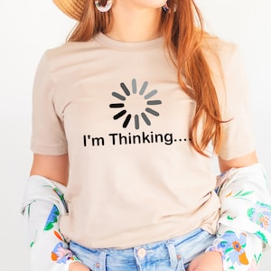 May include: Beige t-shirt with the text "I'm Thinking..." and a loading circle graphic in black and gray. The shirt has short sleeves and a crew neck. The person is wearing a floral patterned jacket.