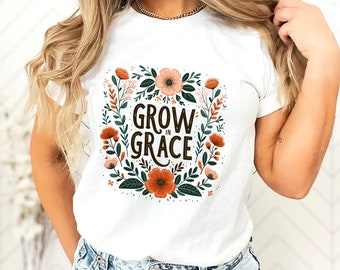May include: White t-shirt featuring a floral design with the words "GROW IN GRACE" in a dark brown font. The floral arrangement includes orange, pink, and green elements. The shirt is made of a soft material.