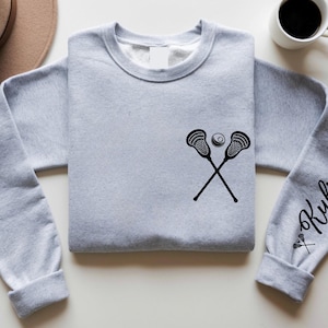 May include: A light gray sweatshirt featuring a black lacrosse stick and ball graphic. The sleeve has the name "Kylie" in a script font. A hat and a cup of coffee are in the background.