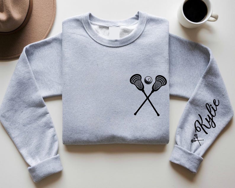 May include: A light gray sweatshirt featuring a black lacrosse stick and ball graphic. The sleeve has the name "Kylie" in a script font. A hat and a cup of coffee are in the background.