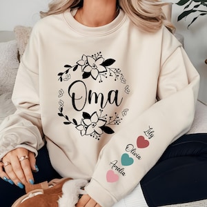 May include: A cream-colored sweatshirt with a floral wreath design and the word "Oma" in the center. The sleeve has three pink, teal, and red hearts with the names Amelia, Olivia, and Lily written next to them.