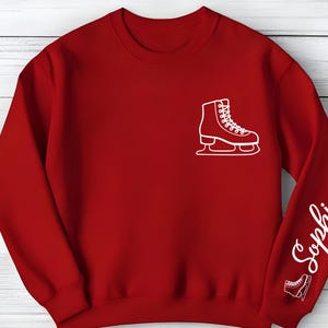 May include: A red sweatshirt featuring a white outline of an ice skate on the chest and the name "Sophia" in white script on the sleeve. A smaller ice skate outline is near the cuff. The sweatshirt is made of a soft material.
